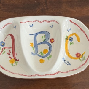 May include: Oval, three-section serving dish with the letters A, B, and C painted in red, blue, and yellow. The dish is white with a red border and floral accents. A decorative piece for serving snacks or appetizers.