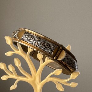 May include: A hinged bangle bracelet featuring a two-tone design with a metallic gold finish and silver-toned panels. The silver sections are decorated with intricate, embossed floral and leaf-like patterns, creating a textured appearance.