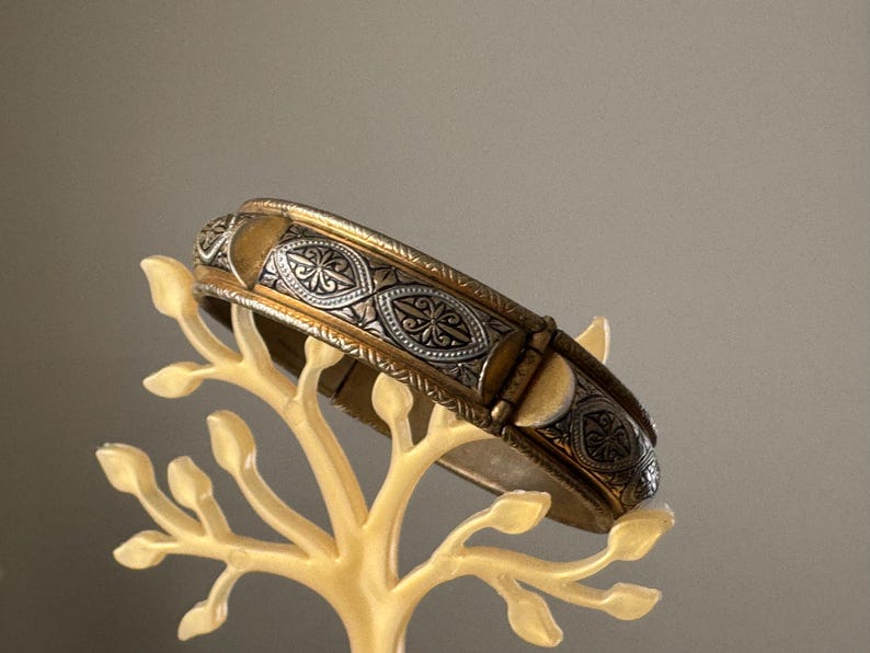 May include: A hinged bangle bracelet featuring a two-tone design with a metallic gold finish and silver-toned panels. The silver sections are decorated with intricate, embossed floral and leaf-like patterns, creating a textured appearance.