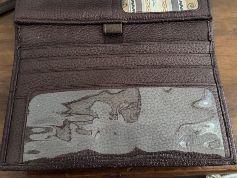 May include: A dark brown textured leather wallet featuring multiple horizontal card slots, a central rectangular clear plastic ID window, and a small top compartment with a patterned fabric lining visible at the upper edge of the open interior.