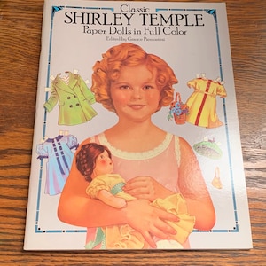 May include: A vintage "Classic Shirley Temple Paper Dolls in Full Color" book. The cover features a portrait of Shirley Temple holding a doll, surrounded by paper doll clothing. The book is on a wooden surface.
