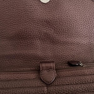 May include: A dark brown textured leather wallet featuring a flap closure with a silver-toned snap button. The interior includes a zippered compartment with a heart-shaped leather pull tab and multiple horizontal card slots along the front panel.