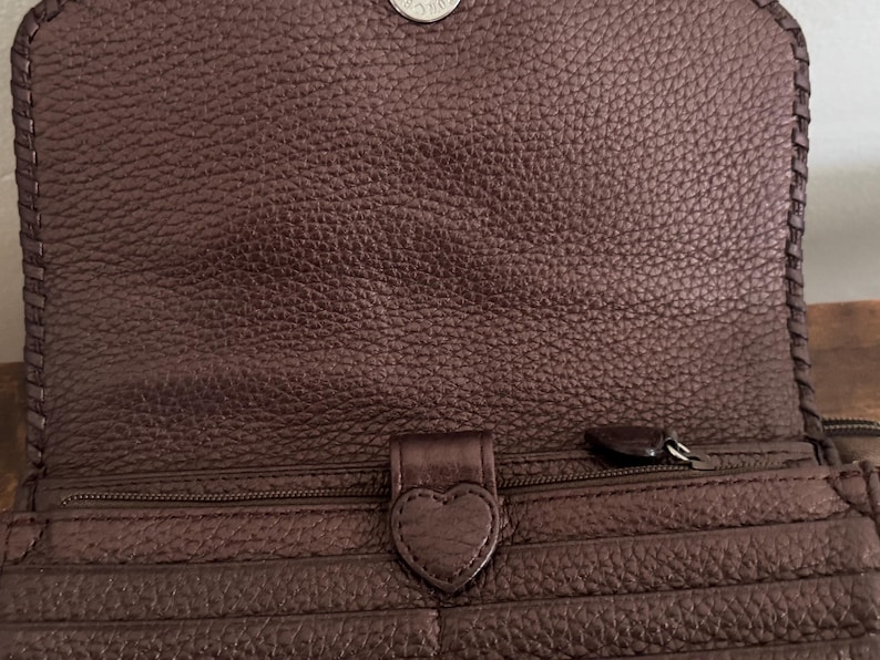 May include: A dark brown textured leather wallet featuring a flap closure with a silver-toned snap button. The interior includes a zippered compartment with a heart-shaped leather pull tab and multiple horizontal card slots along the front panel.