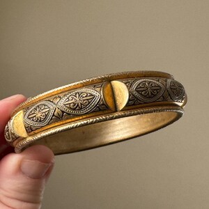 May include: A gold-toned bangle bracelet featuring a central band with alternating smooth gold semi-circles and dark, recessed panels decorated with intricate, engraved floral and leaf patterns, finished with textured, beaded edges.