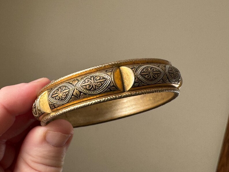 May include: A gold-toned bangle bracelet featuring a central band with alternating smooth gold semi-circles and dark, recessed panels decorated with intricate, engraved floral and leaf patterns, finished with textured, beaded edges.