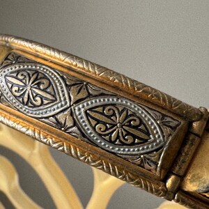 May include: A hinged metal bangle bracelet featuring a two-tone gold and silver finish. The surface is decorated with repeating oval-shaped panels containing intricate floral engravings, bordered by textured, rope-like metal detailing.