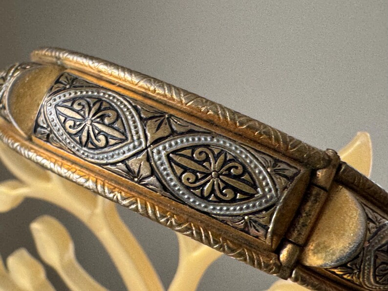 May include: A hinged metal bangle bracelet featuring a two-tone gold and silver finish. The surface is decorated with repeating oval-shaped panels containing intricate floral engravings, bordered by textured, rope-like metal detailing.
