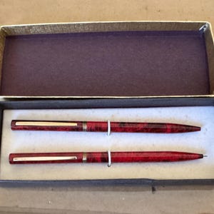 May include: A set of two red and gold pens in a presentation box. The pens have a marbled pattern and gold accents. The box is lined with purple fabric and has a gold trim. The pens are displayed on a white surface.