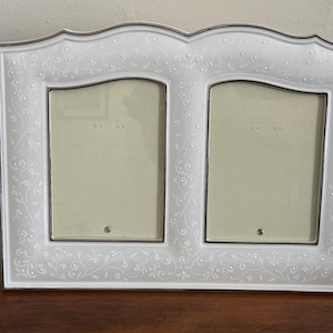 May include: A white double picture frame with a silver trim. The frame has a decorative floral pattern and holds two rectangular photos. The frame is sitting on a dark wooden surface.