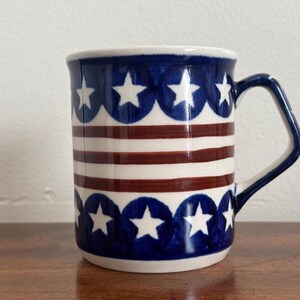 May include: A ceramic mug featuring a patriotic design with a band of white stars on a blue background at the top and bottom, separated by horizontal red and white stripes in the center. The mug has a single handle and a smooth, glossy finish.
