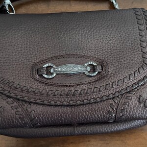 May include: A dark brown pebbled leather crossbody bag featuring a flap closure with decorative whipstitch detailing along the edges. The center of the flap is adorned with an elongated, silver-toned metal hardware piece with intricate engravings.