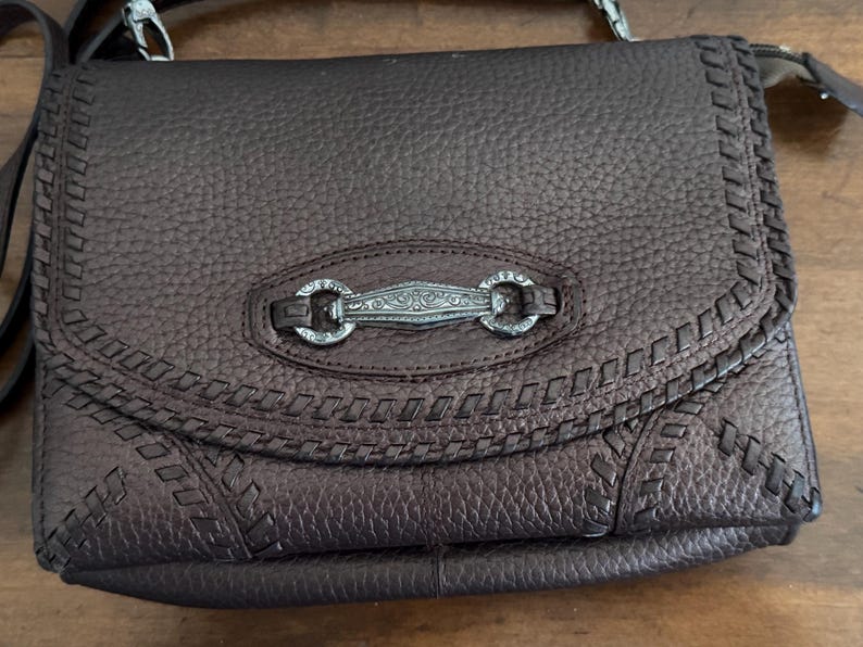 May include: A dark brown pebbled leather crossbody bag featuring a flap closure with decorative whipstitch detailing along the edges. The center of the flap is adorned with an elongated, silver-toned metal hardware piece with intricate engravings.