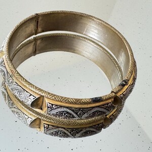 May include: A circular hinged bangle bracelet featuring a two-tone design with alternating gold-colored metal bands and silver-colored panels. The silver panels are decorated with intricate engraved patterns, while the gold bands have a textured finish.