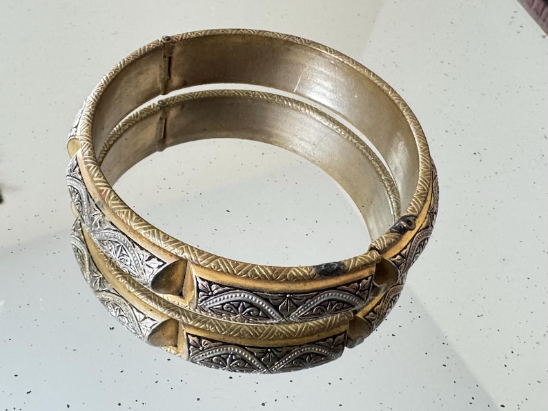 May include: A circular hinged bangle bracelet featuring a two-tone design with alternating gold-colored metal bands and silver-colored panels. The silver panels are decorated with intricate engraved patterns, while the gold bands have a textured finish.