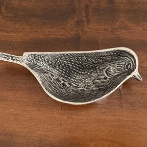 May include: A silver-toned metal trinket dish shaped like a bird in profile. The surface features a detailed, textured pattern mimicking feathers across the body and tail, with a smooth, defined beak and eye area visible on the right side.