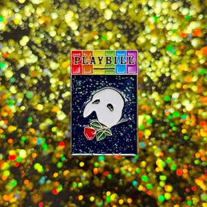Puede incluir: Un pin de esmalte de colores arcoíris con la palabra "PLAYBILL" en letras blancas. El pin presenta una máscara blanca del fantasma de la ópera con una rosa roja sobre un fondo azul oscuro.