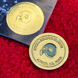 Puede incluir: Pin de color dorado con un diseño de máscara de fantasma negro y verde. El pin dice "Final Performance April 16, 2023".