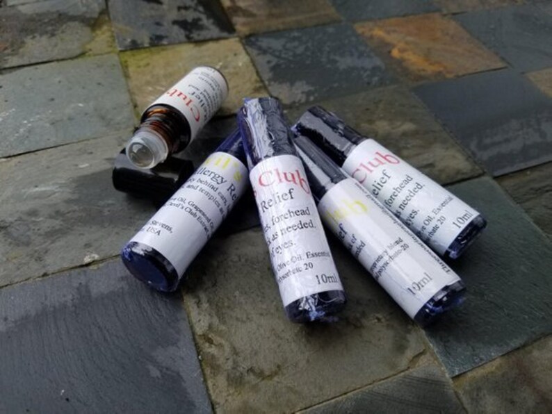 K&ouml;nnte beinhalten: Mehrere kleine, zylindrische Fl&auml;schchen mit &auml;therischen &Ouml;len, wei&szlig;en Etiketten und schwarzen Kappen liegen auf einer strukturierten, dunklen Oberfl&auml;che. Die Etiketten tragen das Wort "Club" in Rot.