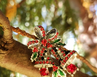 Könnte beinhalten: Silberfarbene Weihnachtsglockenbrosche mit roten und schwarzen Akzenten und grünen Stechpalmenblättern. Die Brosche ist mit roten Edelsteinen besetzt und vor einem verschwommenen Hintergrund aus grünem Laub und einem braunen Ast platziert.