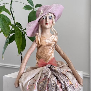 May include: A decorative doll with a porcelain face, painted features, and blonde hair, wearing a wide-brimmed lavender straw hat. The doll has long fabric arms and wears a tan velvet bodice with a pink tulle bow and a patterned skirt with butterflies.