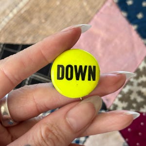 Puede incluir: Un botón redondo y amarillo brillante con la palabra "DOWN" en letras mayúsculas negras y en negrita. El botón se sostiene en una mano, sobre un fondo de retazos de varios colores y texturas.