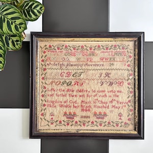 May include: Framed antique sampler with a dark wood frame. The sampler features red and tan cross-stitch embroidery with the alphabet, numbers, and a Bible verse. The text reads "Suffer the little children to come unto me." Dated 1899.