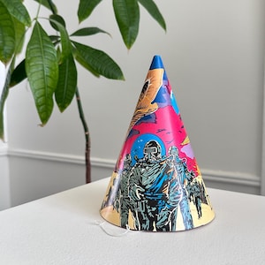May include: A colorful party hat with a cone shape. The hat features a vibrant design with illustrations of figures in protective gear against a backdrop of pink, blue, and yellow hues. A white string is attached at the base.