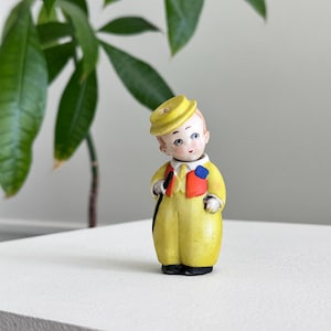 Puede incluir: Una figura vintage de una persona joven con un traje y sombrero amarillos, con un acento rojo y azul en la chaqueta. La figura tiene una cara pintada y está de pie sobre una superficie blanca. Una planta verde está en el fondo.