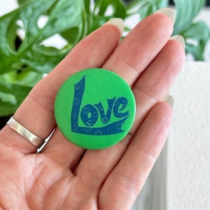 Puede incluir: Un botón verde con la palabra "Love" en azul. El botón es redondo y brillante. La palabra "Love" está estilizada con elementos decorativos.