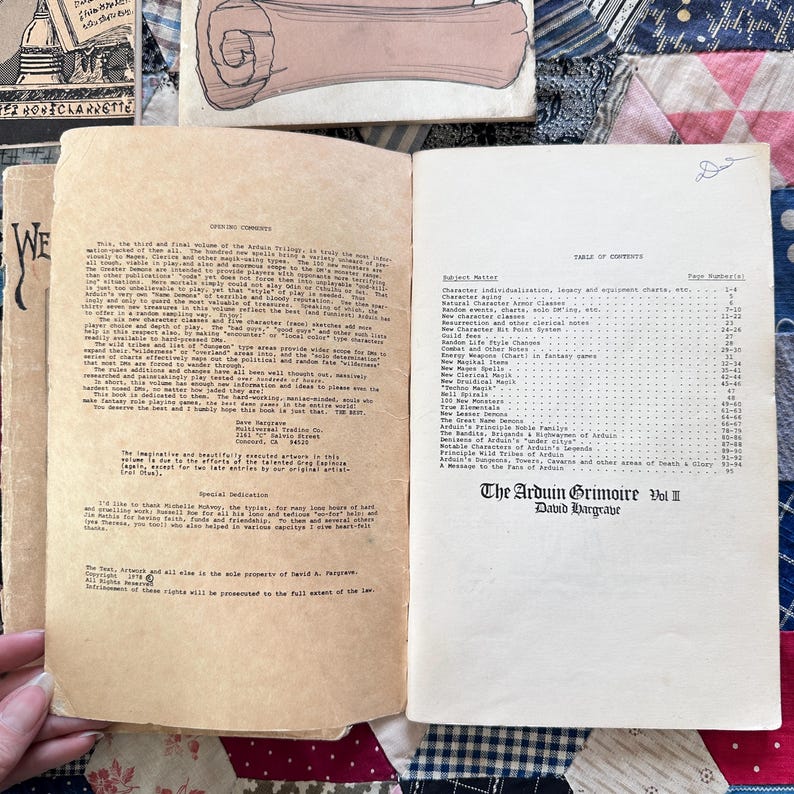 Puede incluir: Un libro abierto con p&aacute;ginas envejecidas y amarillentas. La p&aacute;gina izquierda contiene texto, mientras que la p&aacute;gina derecha muestra una tabla de contenidos. El libro se titula "The Arduin Grimoire Vol II" y est&aacute; fechado. El libro est&aacute; sobre un edred&oacute;n de retazos.