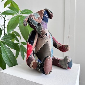 May include: A handmade patchwork dog plush toy. The dog is made from various fabric scraps in a variety of colors, including red, blue, brown, and purple. The toy has a button eye and stitched details. It is sitting on a white surface.