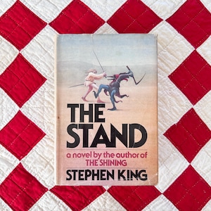 May include: A hardcover book titled The Stand by Stephen King. The cover features a tan background with a central illustration of a person in light clothing fighting a dark, horned creature with a scythe. The text reads The Stand, a novel by the author of The Shining, Stephen King.