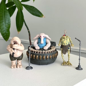 Può includere: Tre action figure aliene di Star Wars. Una figura suona uno strumento a fiato, un'altra è seduta a una tastiera e la terza è in piedi con un microfono. Le figure sono su una superficie bianca con una pianta verde sullo sfondo.