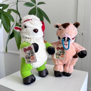 May include: Two plush toys: a sheep and a pig. The sheep is white with red accents and green limbs. The pig is pink with brown spots and a blue nose ring. Both toys have tags that read "Backward Commandos" with a cartoon illustration.