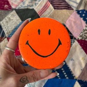 Puede incluir: Un posavasos redondo y naranja con un diseño de carita sonriente negra. El posavasos tiene una textura moteada y se sostiene sobre un fondo de patchwork colorido. La carita sonriente presenta dos puntos negros para los ojos y una línea negra curva para la boca.