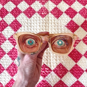 May include: A pair of clear-framed glasses with a unique design. The lenses feature a detailed painting of a human eye with a blue iris. The glasses are held against a red and white checkered quilt.