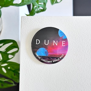 Puede incluir: Un botón redondo con fondo negro y una imagen colorida de un planeta con el texto "DUNE" en letras blancas. El texto "COMING CHRISTMAS WORLD WIDE" está escrito en un círculo alrededor del planeta.