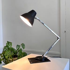 May include: A black desk lamp with a chrome adjustable arm and a square base. The lamp's shade is angled downward, emitting a warm light. The lamp is on a white surface, with a green plant in the background.