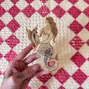 May include: A wooden mermaid comb with a fish. The mermaid has long hair and a floral design on her tail. The fish is black and white. The comb is held against a pink and white quilt.