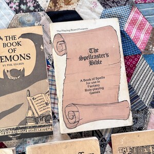 Puede incluir: Dos libros antiguos se muestran sobre un edred&oacute;n de retazos. El libro de la izquierda se titula "The Book of Demons" de Phil Edgren. El libro de la derecha es "The Spellcaster's Bible: A Book of Spells for use in Fantasy Role-playing Games".