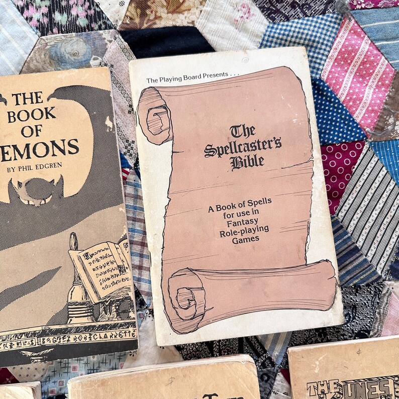 Puede incluir: Dos libros antiguos se muestran sobre un edred&oacute;n de retazos. El libro de la izquierda se titula "The Book of Demons" de Phil Edgren. El libro de la derecha es "The Spellcaster's Bible: A Book of Spells for use in Fantasy Role-playing Games".