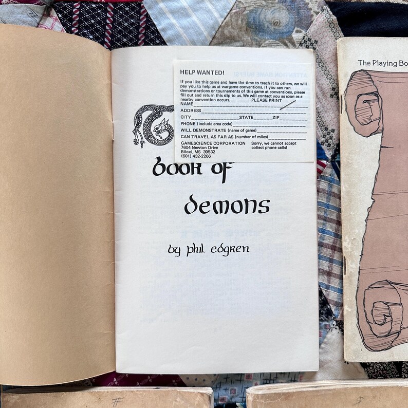 Puede incluir: Libro abierto titulado "Book of Demons" de Phil Eogren. La portada presenta una ilustraci&oacute;n de drag&oacute;n y texto. Se adjunta un formulario "Help Wanted!". El libro est&aacute; sobre una superficie estampada.