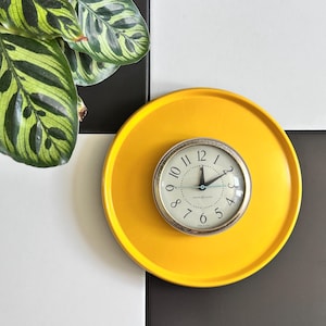 Puede incluir: Una bandeja redonda amarilla con un reloj con borde plateado en el centro. El reloj tiene una esfera blanca con números y manecillas negras. Una planta verde y blanca a rayas está en la esquina superior izquierda. El fondo es a cuadros blancos y negros.