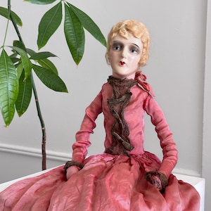 May include: A vintage doll with a painted face, blonde hair, and a pink dress. The doll's dress has long sleeves and a ruffled brown collar. The doll is sitting, and the skirt of the dress is spread out. The doll has a red bow on the back of its head.