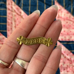K&ouml;nnte beinhalten: Eine kleine, goldfarbene Metallbrosche mit dem Wort "BEBE" auf einem Banner. Die Brosche hat an jedem Ende ein kleeblattf&ouml;rmiges Design. Die Brosche wird in einer Hand vor dem Hintergrund eines rosa-blauen Quilts gehalten.