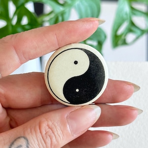 Puede incluir: Un botón redondo con el símbolo del Yin y Yang. El diseño se divide en dos secciones curvas, una negra y otra blanquecina, cada una con un pequeño punto negro. El botón se sostiene entre los dedos.