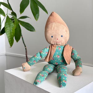 May include: A vintage cloth doll with a tan head and a pointed hat. The doll has blue eyes and a stitched smile. It wears a teal floral print outfit with a tan vest. The doll is sitting on a white surface.