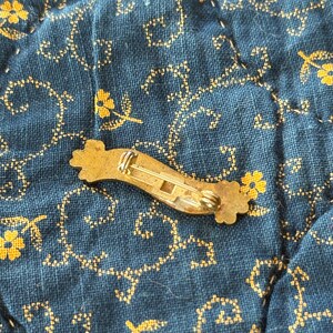 K&ouml;nnte beinhalten: Vintage Brosche aus goldfarbenem Metall mit geschwungenem, band&auml;hnlichem Design. Die Brosche hat florale Akzente an jedem Ende und einen Nadelverschluss auf der R&uuml;ckseite. Die Brosche liegt auf einem blauen Stoff mit gelbem Blumenmuster.