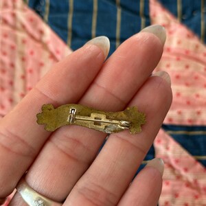 K&ouml;nnte beinhalten: Eine Vintage-Anstecknadel aus Messing mit floralem Design an den Enden. Die Nadel hat einen einfachen Stangen- und Verschlussverschluss. Die Nadel ist leicht gebogen und hat ein angelaufenes Aussehen, was auf das Alter hindeutet.