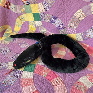 May include: A plush black snake toy with a soft, fuzzy texture. The snake features a red tongue, green eyes with glittery black accents around them, and a blue gem embedded in a black collar-like band around its neck area, coiled on a surface.